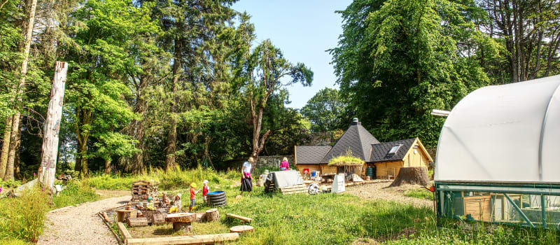 MAKI Pups outdoor nursery site with children playing and a polytunnel, hobbit house and woodland in background